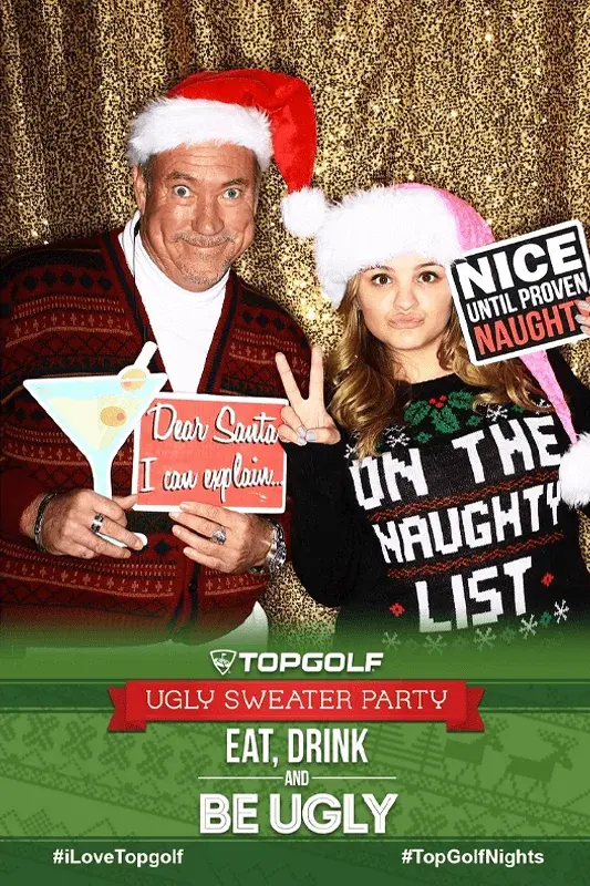 Two people in holiday sweaters and Santa hats posing with festive signs at a photo booth.